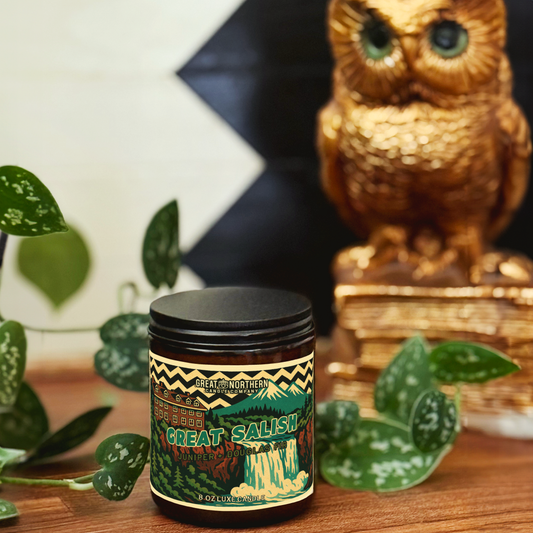 Candle jar with 'Great Salish' branding on a wooden surface with plants and an owl figurine in the background.