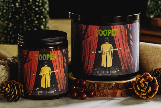 Two Container Candles, in Black, surrounded by nature elements, pinecones, berries, wood rounds. With a colorful Agent Cooper Label.